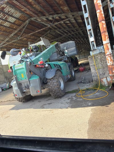 This image, captured in Bazan, Bulgaria, depicts an industrial or agricultural setting, likely inside or at the entrance of a large workshop or shed. The main subject is a green and grey Kramer telehandler or compact loader, prominently parked on a concrete surface. The vehicle features large, rugged tires and has "KRAMER" clearly visible on its side in white lettering, with smaller text above it appearing to read "APOTHEUM" and a yellow warning symbol. Another piece of green machinery, possibly a tractor or additional equipment, is partially visible behind the Kramer, bearing the text "P 12" and what looks like part of "CLAAS".

Overhead, a network of aged, rusted metal pipes and grey support structures extends across the ceiling, suggesting a processing or storage facility. A bright yellow hose is coiled on the ground, extending from a red air compressor positioned behind the Kramer vehicle. The scene is illuminated by strong natural daylight, indicating it is daytime, which casts distinct shadows on the concrete floor. The ground itself is a mix of concrete and what appears to be a coarser, lighter-colored aggregate. No individuals are present in the image.