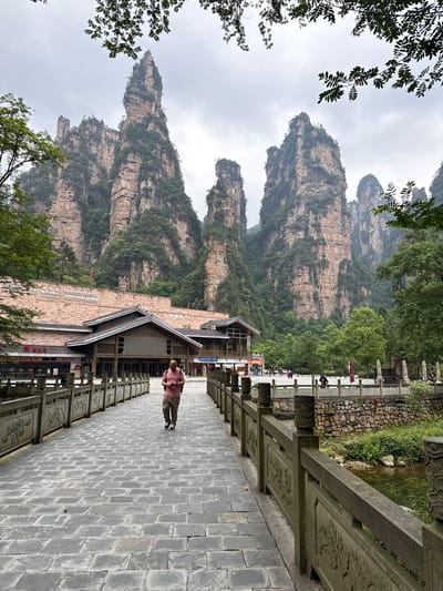 A landscape photograph captures a paved walkway lined with an ornate, stone railing, with a person in a pink shirt walking away from the viewer. The background features tall, rocky mountains covered with sparse vegetation, receding into the overcast sky. A building with a traditional-style roof is situated adjacent to the walkway. The scene appears to be located in or near a developed area in Xiehe, China.
