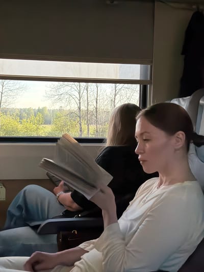 A photograph taken on a train in Ushaki, Russia, shows two individuals seated by a window. The person in the foreground has dark hair pulled back in a bun and is wearing a white top. They are reading a document. The second individual, seated next to them, has blond hair and is wearing a black shirt and blue jeans. A brown bag rests next to them. Through the window, a view of trees and a partially-obscured landscape are visible.