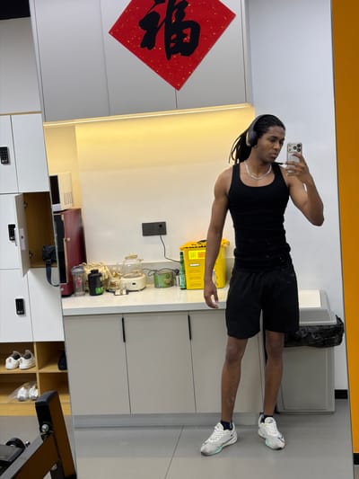 A person stands in front of a mirror, taking a selfie with a smartphone. The person is wearing a black tank top, black shorts, headphones, and white sneakers. The environment appears to be an indoor space, possibly a home or gym. The background includes white cabinets, a red decorative square, and a trash can. The scene is potentially located in the Binjiang District, China, as suggested by the background.