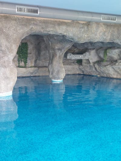 The image shows a swimming pool in Ognyanovo, Bulgaria, with a rock-like structure resembling a cave. The pool is filled with clear blue water, and the edges are lined with small mosaic tiles. Above the pool, a grey air conditioning duct runs along the ceiling, with a vent visible. Lush green ivy is growing on some parts of the rock formation. The overall scene depicts an indoor or partially covered swimming area with a naturalistic design.