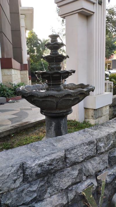 The image shows a multi-tiered fountain made of dark stone. Water flows from the top tier into lower basins, with several streams cascading over the edges. The fountain sits on a pedestal, which is in turn placed on a paved area with a patterned design. To the left of the fountain, a building with brown and beige exterior panels is visible, along with some green bushes and a pair of sandals on the ground. To the right of the fountain, there is a white architectural column and a wall constructed of rough, grey stones. Some greenery, including trees and grass, is present in the background. A metallic object, possibly part of a sign or decoration, is partially visible in the lower right corner of the image, resting on the stone wall. The scene appears to be outdoors, possibly in a courtyard or garden area. The location is identified as Sidodadi, Indonesia.