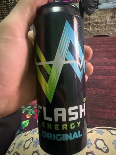 A human hand with light skin tone is holding a black "Flash Energy Original" beverage can in an indoor setting in Qoraobdol QFY, Uzbekistan. The can features a green stylized "V" and a blue stylized "A" on its label, with "UP" printed above "FLASH". The hand grips the left side of the can, with fingers partially visible. Below the can, a light-colored pillow or blanket patterned with white penguins and blue snowflakes is visible, alongside a dark red fabric with a floral pattern. The background is out of focus, revealing a dark reddish-brown surface, likely a floor or rug, and a dark piece of furniture or object.