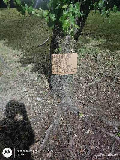 This low-light image, captured on October 30, 2023, at 4:06 AM, depicts a natural outdoor environment described as being in Tulsa, United States. The primary subject is a tree with a rough, textured bark and visible root system extending into the surrounding dirt and scattered debris. Green foliage is visible in the upper portion of the tree.

Affixed to the trunk of the tree, approximately mid-frame, is a rectangular piece of light brown cardboard serving as a handmade sign. The sign features handwritten text in dark marker, oriented vertically, which reads: "STOP THE FBI TERRORIST WATCHLIST GANGSTALKING CHILDREN ON IT".

The ground cover consists of sparse green grass in the background, transitioning to exposed dirt and small stones around the tree's base and roots in the foreground. A distinct, dark human shadow, likely belonging to the photographer, is cast on the lower-left ground area. The illumination suggests artificial light source, possibly from the camera's flash or a nearby street lamp, creating bright spots on the foliage and ground.