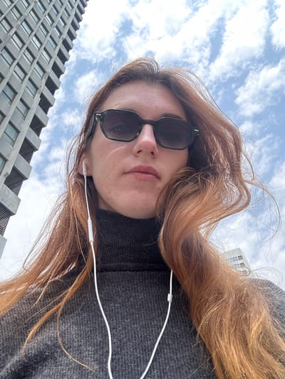 A person with red hair and sunglasses, wearing a gray sweater and earphones, is looking up at the camera. The background consists of a partly cloudy sky and the side of a modern building with a regular grid of windows. The location is the city of Huangdao District, China.