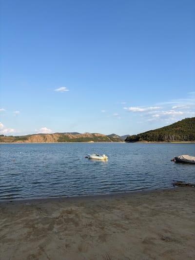 An eye-level, vertical, medium shot captures a serene lake scene under a bright blue sky dotted with wispy clouds. The foreground features a sandy beach with tire tracks. The middle ground is dominated by a wide expanse of calm blue water, rippled by a gentle breeze. Two small boats are visible; one is a white motorboat with a yellow trim, anchored in the center of the lake, and another is partially covered by a grey tarp, moored to the right. In the background, rolling hills covered with dry brush and scattered green trees rise from the water's edge, with more densely forested hills on the right. The overall atmosphere is peaceful and natural.