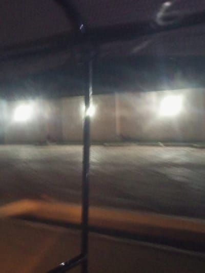 The media file depicts an indoor parking area, likely at night, given the artificial lighting. The scene is dimly lit, with two prominent rectangular light sources casting a bright glow on a light-colored wall. The floor of the parking area appears to be asphalt or concrete. In the foreground, a dark, vertical pole is visible, suggesting it is part of the parking structure or a barrier. The overall mood is somewhat desolate and industrial. There are no people or vehicles visible in the frame. The lighting creates a diffuse, almost hazy atmosphere, and the image has a slightly grainy quality. The lack of natural light and the presence of artificial lighting strongly indicate it is nighttime. The location context is Baccure Kofare, Nigeria. There is no visible text in the image.