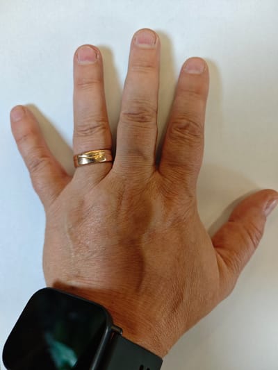 A person's hand with five fingers is shown with a gold ring on the third finger from the left and a black smartwatch on the wrist. The hand is resting on a white surface.