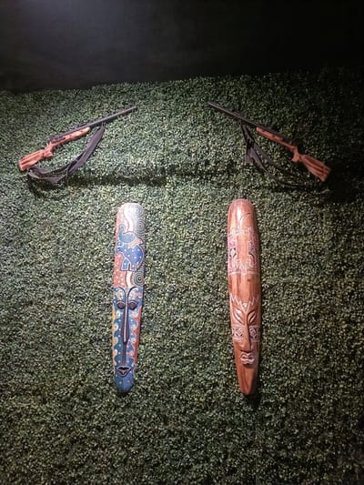 The image shows two wooden masks displayed on a wall covered in dense green foliage. Above the masks, two rifles with slings are mounted on the wall. The lighting is dim, casting shadows and highlighting the textures of the foliage and the masks. One mask is on the left, intricately painted with blue, yellow, and white patterns, featuring a central face with dark eyes and a patterned headdress. The other mask is on the right, made of natural wood with carved patterns, including what appears to be animal motifs and a human-like face. The rifles appear to be decorative. The scene suggests a curated display, possibly in a themed room or an exhibition, aiming for a rustic or perhaps even a slightly menacing aesthetic given the presence of the rifles. The foliage might be artificial, creating a textured backdrop. No text is visible in the image. The overall mood is mysterious and visually striking due to the contrast between the organic foliage, the carved masks, and the metallic rifles. The specific location cues for Kurnool, India, are not directly evident from the visual content, but the style of the masks could potentially be indicative of indigenous art from that region or similar regions in South Asia. The time of day is likely evening or night, given the artificial lighting.