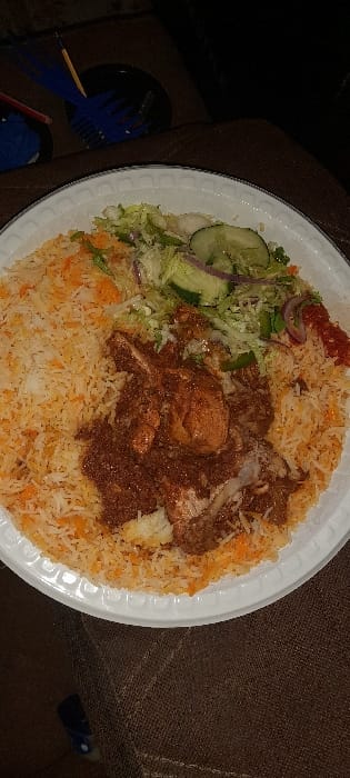 A single meal is presented on a white, disposable, round plate with a decorative rim. The plate holds a large portion of two-toned rice, featuring white and orange-yellow grains, covering the majority of its surface. Centrally placed over the rice is a dark reddish-brown stew containing several pieces of chicken, with visible bone structures. Adjacent to the stew and rice, in the upper right quadrant, is a fresh salad composed of shredded green lettuce, sliced cucumbers, and red onion slivers. A small dollop of bright red, textured chili relish is present on the far right edge of the plate. In the dark background, a dark plate or bowl contains two blue plastic forks and a wooden skewer. The entire assembly rests on a dark brown, textured fabric surface. This scene is located in Ziwa La Ng'ombe ward, Kenya.