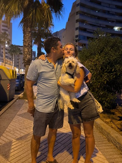A man in a blue denim shirt and grey shorts kisses a woman on the cheek. The woman, wearing a light blue shirt and denim shorts, holds a small, fluffy white dog. The dog has its mouth open, as if panting or barking. They are standing on a sidewalk made of light-colored rectangular pavers, with a curb to their right. Behind them are palm trees, a building with balconies, and a street with a parked car. To the left, there are two large, grey trash bins with yellow lids. The scene is lit by artificial lights, casting long shadows on the sidewalk. The overall impression is a casual moment captured outdoors at dusk or early evening in Calp, Spain.