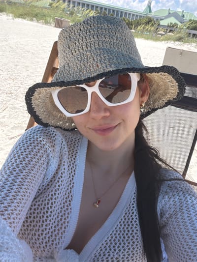 A woman wearing a straw hat and sunglasses poses for a selfie on Hilton Head Island, South Carolina, United States of America. She is seated on a beach chair, with a white, crocheted cover-up over her shoulders and a necklace with a red pendant. The background shows a sandy beach, dunes with sea grass, and a large resort building with a green roof. The sky is partly cloudy.