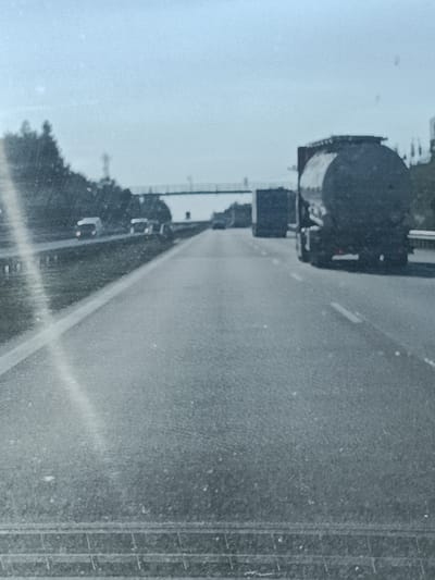 The image shows a highway scene from the perspective of a vehicle's dashboard. The road is asphalt with white lane markings visible. On the right side of the highway, there is a large tanker truck. Further ahead, another truck is partially visible, and beyond that, two vans are driving in the distance. On the left side of the highway, trees and vegetation line the road. A pedestrian bridge spans across the highway in the distance. The sky is overcast, suggesting a cloudy day. The overall scene depicts traffic on a highway. There is no visible text in the image.