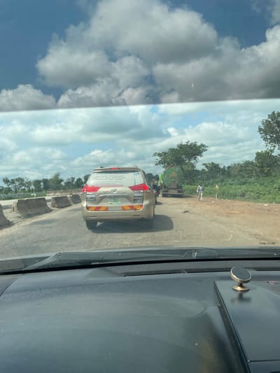 The image shows a view from inside a vehicle, looking out at a road scene in Aciol, Nigeria. The perspective is from the passenger seat, with part of the car's interior, including a black dashboard and a circular magnetic phone holder, visible on the right side of the frame. The main focus is the road ahead, where a light-colored SUV is in the foreground, followed by a green tanker truck. In the distance, a person in white clothing is standing near the side of the road. The setting appears to be rural, with dense green vegetation and trees lining the right side of the road. The sky is partly cloudy, with patches of blue sky visible between large, white clouds, suggesting it is daytime. The road surface is asphalt, with some rough patches and debris visible, indicating potentially developing infrastructure or maintenance work.

The rear of the SUV has several stickers and text on it. Visible text includes company names like "DUNAMIS" repeated multiple times, and what appears to be a list of locations: "UGU PORT HARCOURT", "ABUJA", "REIN UTD LAGOS WARRI BATELSA", "CALABAR JOS KAND KADUNA". A phone number is also visible: "08100079208922658295". The tanker truck has some text in yellow lettering on its side, though it is mostly obscured. The overall impression is a snapshot of daily life and transport on a Nigerian road.