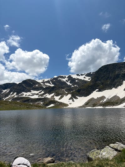 The image depicts a serene alpine lake in Panichishte, Bulgaria, situated at the base of snow-capped mountains under a clear blue sky dotted with fluffy white clouds. The water of the lake is dark and slightly rippled, reflecting the surrounding landscape. The mountains in the background are rugged and streaked with patches of snow, indicating a high-altitude environment. Green vegetation covers the lower slopes of the mountains and the foreground. In the lower right corner, rocks and grass are visible, with a portion of a white and black shoe in the bottom left corner. The scene conveys a sense of tranquility and natural beauty.