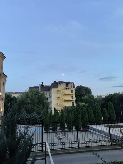 The image shows an outdoor scene in Veshenskaya, Russia, featuring buildings, trees, and a fence. In the upper portion of the image, a pale, full moon is visible against a twilight sky with scattered clouds. The sky transitions from a lighter blue near the horizon to a deeper blue overhead.
Dominating the mid-ground are several buildings. To the left, a portion of a building with beige stone facade and arched windows is visible. Adjacent to it is a multi-story yellow building with dark brown roof. This building has a prominent external yellow staircase winding up its side, with balconies on each level. Several air conditioning units are mounted on the exterior walls of this yellow building. To the right of the yellow building, a taller, darker building with a brown roof and visible air conditioning units is partially obscured by trees.
The foreground is occupied by a black metal fence with an intricate circular design and vertical bars. Behind the fence, a paved area, possibly a courtyard or recreational space, is visible. Several green coniferous trees, some tall and slender and others more rounded, are arranged in rows behind the fence. More mature deciduous trees with dark green foliage are situated further back, behind the fence and in front of the buildings. A dark green, bushy evergreen shrub is in the immediate foreground on the left, partially obscuring the fence. The ground in front of the fence appears to be paved with asphalt.