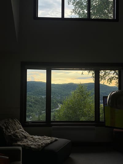 This image depicts an indoor scene in Mont-Tremblant, Canada, featuring a large window overlooking a heavily wooded landscape at dusk. The window is divided into multiple panes, revealing rolling hills covered in lush green trees. The sky above is a mix of soft clouds and the warm glow of sunset, with hues of orange and yellow.
In the foreground, to the left, a dark chaise lounge is draped with a thick, textured faux fur throw and a matching pillow. The lounge is positioned to face the window, suggesting a comfortable viewing spot. To the right of the window, a television screen is partially visible, displaying a bright, possibly green, image. The overall ambiance is serene and tranquil, with the interior space cast in shadow by the dimming natural light. The architecture suggests a modern home or chalet, likely designed to maximize the scenic views of the Mont-Tremblant region.