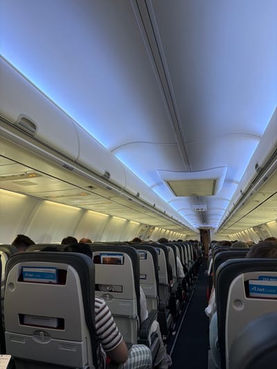The image shows the interior of a commercial airplane cabin, viewed from the perspective of a passenger seated on the right side of the aisle, looking forward. Rows of seats with dark gray upholstery and black headrests extend down the length of the cabin. Each seatback has a tray table, a seat pocket containing literature, and a screen. Overhead compartments are visible along the top of the cabin, with soft blue lighting emanating from the ceiling. Passengers are seated in many of the seats, their heads and shoulders visible. Some passengers are wearing casual clothing. An "AJet CAFE" card is visible in a seat pocket. A sign with Turkish text, "CİKİŞ EXIT," is visible in the distance towards the rear of the cabin. The overall environment is that of a typical aircraft interior during boarding or a flight. There is no visible indication of the specific location within the city of Pendik, Turkey, within the image itself.