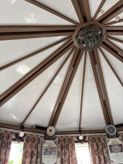 The image shows the interior of a round structure, possibly a gazebo or a covered seating area, with a white, translucent roof supported by brown wooden beams radiating from a central point. A multi-tiered, crystal chandelier hangs from the center of the roof. Along the perimeter, decorative curtains with a brown and beige patterned fabric are visible, along with framed posters depicting food dishes, one of which is labeled "Дагестанские Курзе" and another "Пельмени СССР Узбекские". Several small circular decorative plates are mounted on the beams. Two windows are partially visible on the left and right sides, showing greenery outside. The overall impression is of a food establishment or a dining area with a rustic and decorative ambiance. The location is identified as Русский Турек, Russia.