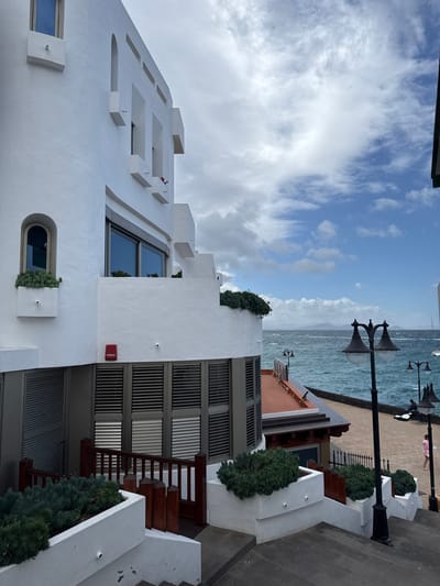 The image depicts a multi-story white stucco building with Mediterranean architectural elements situated on a paved pathway leading to a waterfront promenade in Playa Blanca, Spain. The prominent building on the left features various rectangular and arched windows, some with integrated planter boxes containing green succulent-type foliage. A red rectangular fire alarm pull station labeled "FUEGO" is mounted on a lower white wall, above multiple closed horizontal slatted shutters. Several small, white cylindrical security cameras are visible on the building's exterior.
Below the main building, dark grey paved steps descend towards a lower level with dark brown wooden railings and additional white planter boxes filled with green foliage. To the right, beyond the steps, lies a paved promenade bordering a turbulent, dark turquoise sea with whitecaps. Distant landmasses are visible on the horizon under a partially cloudy blue sky.
Along the promenade, multiple black, ornate streetlamps with two globes each are present. One individual, dressed in a light-colored top and shorts, is walking away from the viewer on the promenade. Another individual is seated further down the promenade, partially obscured by a streetlamp. At least two small white boats are visible in the distant sea.