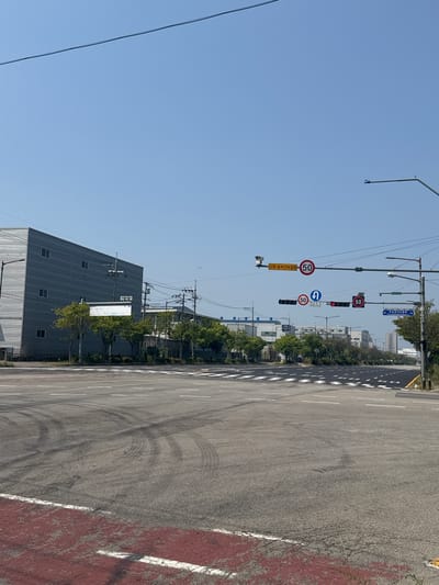 The image shows an intersection in Pyeongtaek-si, South Korea, under a clear blue sky. The intersection is paved with asphalt and has white lane markings and a red-colored pedestrian crossing. Tire marks are visible on the road surface, indicating recent activity.
On the right side of the intersection, a traffic light pole stands with several signs and traffic signals attached. These include speed limit signs indicating "50" in red circles, a camera, and a blue sign with Korean text.
In the background, there are several industrial buildings with gray exteriors and rows of windows. Trees line the area between the road and the buildings. A power line stretches across the top of the image. The overall scene depicts a quiet, industrial area.