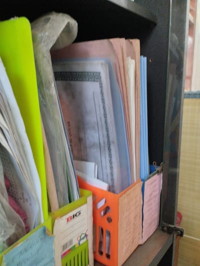 The image depicts a close-up, eye-level view of a shelf containing various papers and documents, likely in an office or educational setting. The objects are organized in a binder or container. On the left, a bright green binder is prominent, filled with papers, some of which appear to be in plastic sleeves. To the right of the green binder, a stack of documents is visible, some with light-colored covers. Further to the right, a bright orange container holds additional papers. On the right side of the shelf, a dark vertical element, possibly a cabinet or bookshelf edge, frames the scene. The background shows a textured surface, likely a wall, and a hint of a window with blinds. There are no people or animals visible in the image, and no specific actions are being performed. The overall impression is one of an organized but possibly cluttered collection of documents. The information about the location being Tanjungsari, Indonesia, is not derivable from the visual content of the image itself.