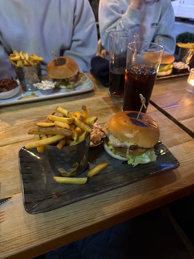 The image shows a table in a dimly lit setting, possibly a restaurant or pub. On the table, there are two plates of food. Each plate contains a burger and a serving of fries presented in a decorative metal holder, along with what appears to be a side of coleslaw. A skewer holds the burger together. Next to one of the plates are two glasses filled with a dark liquid, likely a beverage. Two people are partially visible in the image, one wearing a light grey top. The table itself has a wooden surface.
Given that the user is asking for the location in Windermere, United Kingdom, I can only assume that the location of the food and people are somewhere in Windermere, United Kingdom. I don't have any other information to indicate a specific restaurant name, or location.