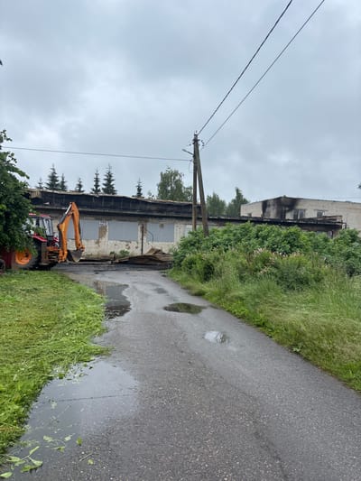 A red tractor with an orange excavator attachment is parked on a wet asphalt road in front of a damaged, single-story brick building. The building appears to be under demolition or has been damaged by fire, with a charred roofline and boarded-up windows. Debris, including corrugated metal sheets, is scattered in front of the building. Overgrown green grass and bushes line the sides of the road and the building. The sky is overcast and cloudy. A wooden utility pole with wires runs across the top of the image. The location is the village of Ushaki, Russia.