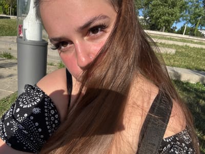 A young woman with long brown hair and large false eyelashes is positioned in the foreground of this outdoor image, her face turned towards the camera. She is wearing a black off-the-shoulder top with a white floral pattern. A silver nose stud is visible on her right nostril. The sunlight casts shadows across her face and hair. In the background, a grey pole with a red sticker is visible to the left, along with tiered grassy areas and trees, suggesting a public park or outdoor recreational space. The setting appears to be in the city of Weil am Rhein, Germany.