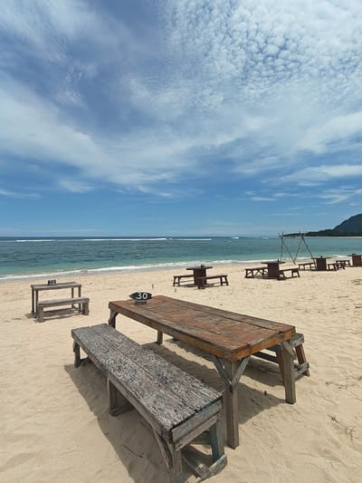 A photograph shows a beach scene, most likely in Monikeun, Indonesia. The setting includes a sandy beach leading to an ocean with waves visible in the distance. The sky is bright with a mix of blue and white clouds. Scattered across the beach are wooden tables and benches. Closer to the camera is a large wooden table with a number "30" displayed on top of it, accompanied by a long, weathered wooden bench. In the background, additional tables and benches are visible, including one with the number "22". A structure resembling a swing set is also present further down the beach. The photograph captures a clear, sunny day at the beach.
