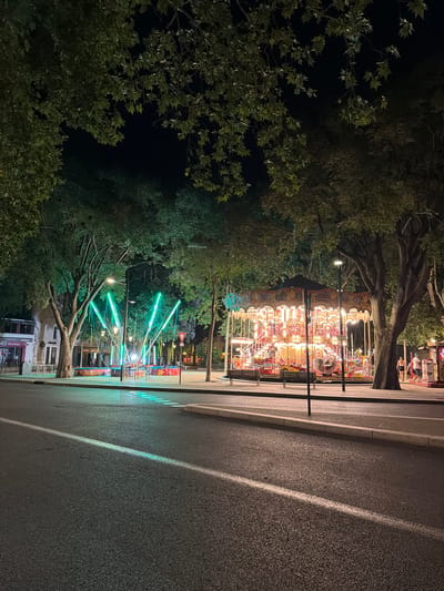 This is a nighttime photograph of a street scene in the city of Nîmes, France, specifically on the Esplanade Charles de Gaulle. A brightly lit carousel, adorned with numerous bulbs and decorative elements, is the central focus of the image. To the left of the carousel, a bungee jumping attraction with bright green lights is visible. Trees with dark green foliage line the street, their branches reaching overhead and creating a canopy. The street itself is asphalt, with a white line marking a lane. In the background, buildings are partially visible, and a few pedestrians can be seen walking along the sidewalk near the carousel. The overall atmosphere is one of a lively urban environment at night, likely a fairground or festival.