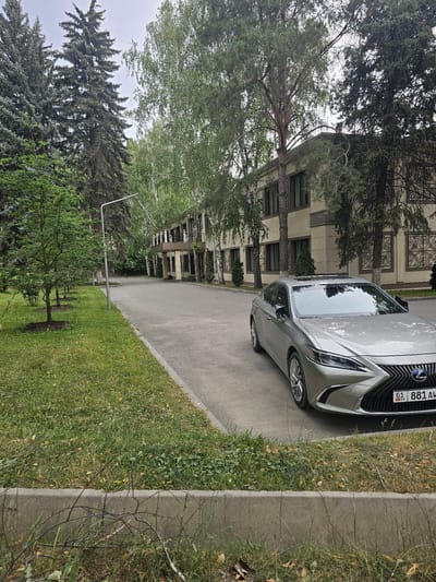 The image displays a silver Lexus sedan parked on an asphalt road next to a grassy area with a concrete curb. The car is facing towards a building in the background, which appears to be a multi-story structure with a light-colored facade and dark-framed windows. Trees with green foliage are scattered throughout the scene, with some large trees casting shadows on the road and grass. A street light pole is visible in the middle ground. The sky is overcast, suggesting a cloudy day. The license plate of the car is partially visible and displays the text "01 KG 881 AW". The overall scene depicts a quiet, possibly residential or institutional setting in Chong-Aryk, Kyrgyzstan, with the car parked and no people visible. The image appears to have been taken during daylight hours.