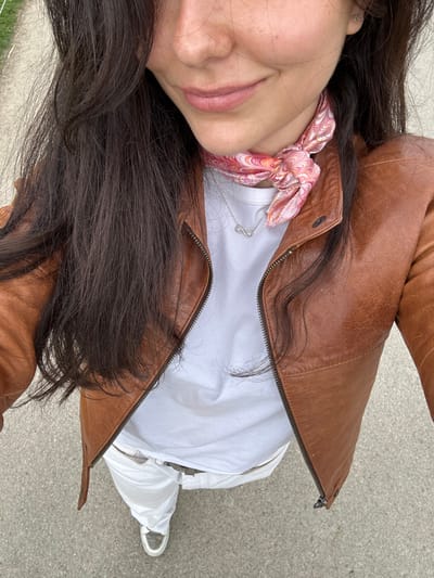 A young woman with long dark brown hair is wearing a brown leather jacket, a white t-shirt, white pants, and a pink and orange bandana. She is wearing a silver infinity necklace and has a faint smile on her face. The woman is standing on a gray paved surface, possibly a sidewalk in the town of Chambord, France, located at latitude: 47.615, longitude: 1.518. She is taking a selfie. The photo appears to be taken from a slightly downward angle. The woman is facing the camera. The background is blurry and contains a few green patches, possibly grass.