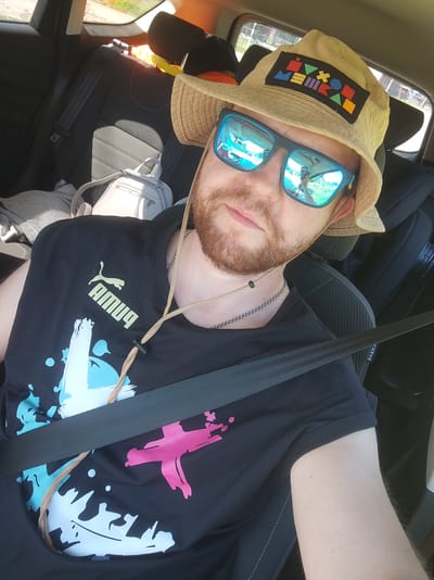 A man wearing a bucket hat and sunglasses is sitting in the passenger seat of a car, looking at the camera. The hat has a colorful patch on the front with geometric shapes. He is wearing a black Puma t-shirt with a white and blue splatter design on the left side and a pink splatter design on the right side. A car seatbelt is across his chest. The background shows the interior of the car, including the headrest and seats, as well as the reflection of the outside world in his sunglasses. There is a white backpack on the seat behind him. The scene is indoors, likely in a car, and there is no specific information about its location within Zashchita, Russia.