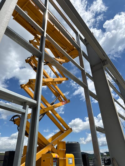 This low-angle, full-shot photograph, taken outdoors, captures a construction site in Skrzydlna, Poland. The foreground and midground are dominated by the yellow scissor lift, its intricate metal support structure extended upwards, reaching towards the sky. The lift is positioned between several large, grey steel beams that form part of an unfinished building structure. Above and behind the construction elements, a bright blue sky is visible, dotted with scattered white clouds. In the background, a slight hazy outline of distant hills and greenery can be observed. The scene suggests ongoing construction work under fair weather conditions.