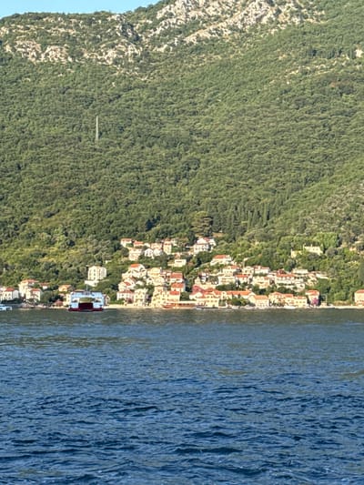 This is a distant, wide shot of a coastal town nestled at the base of a densely forested mountain range. The water in the foreground is a deep blue, with gentle waves suggesting calm conditions. Along the shoreline, a cluster of white buildings with red-tiled roofs forms a picturesque village. A prominent ferry, white with a red upper deck, is docked near the shore, indicating a transportation hub. The mountain slope behind the village is covered in thick green foliage, with occasional rocky outcrops and a solitary transmission tower visible near the summit. The sky is a clear, pale blue. The location is Jošice, Montenegro.