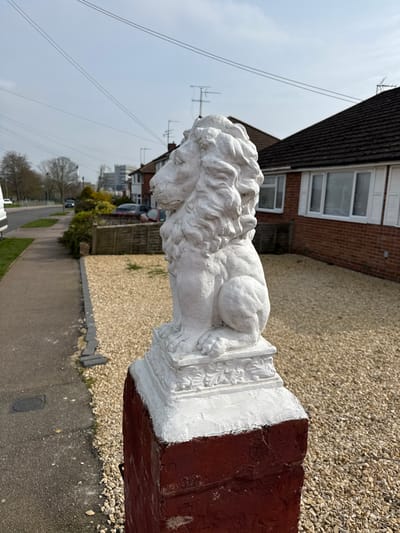 The scene, located in Cherwell District, United Kingdom, depicts an outdoor residential setting. A weathered white statue of a sitting lion, facing left, occupies the central right foreground, positioned on a decorative white plinth atop a reddish-brown brick pillar. To the immediate right of the pillar is a light-colored gravel-covered yard. To the left, a dark asphalt footpath runs parallel to the gravel area. Beyond the footpath, a grassy verge borders a paved road. A white van is partially visible parked on the far left, along the road.
In the midground, to the right of the lion, a single-story house constructed of reddish-brown brick with a dark tiled roof and white-framed windows, some featuring white shutters, is visible. Beyond the road, a line of similar residential houses, characterized by dark roofs, can be seen. Trees, mostly bare of foliage, are interspersed among the houses and along the roadside. In the distant background, a multi-story building with a light-colored facade is discernible. The sky is clear blue with few clouds, and multiple overhead utility wires traverse the upper portion of the image. Several television antennas are visible on the rooftops of the houses. No individuals, actions, or interactions are present in the image.