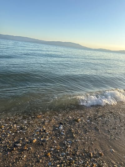 The image captures a serene view of Ofrynio Beach, Greece, at what appears to be dawn or dusk. A gentle wave is rolling onto a pebble-strewn shore, its white foam catching the soft light. The water is a clear, tranquil blue, stretching towards a distant, hazy coastline of mountains under a pale blue sky. The beach itself is covered in a mosaic of smooth, rounded pebbles of various sizes and colors, interspersed with sand. The overall impression is one of peaceful natural beauty.
