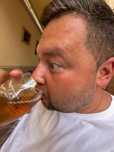 A close-up, eye-level shot shows a man in his late 30s or early 40s drinking from a clear glass of beer. He has fair skin, brown hair with visible grey streaks, and a beard and mustache. He is wearing a white t-shirt. The man is outdoors, with a yellow building and a small window visible in the blurred background. The scene appears to be a public outdoor seating area, possibly a restaurant or bar, in the city of Sant Feliu de Guíxols, Spain. The glass has text printed on it in white lettering. The man is holding the glass with his left hand and tilting it to drink. His eyes are open and he is looking to his right.