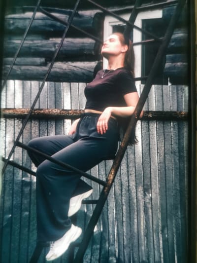This media file features a young woman posed on an outdoor metal ladder. She is wearing a dark, form-fitting t-shirt and dark, loose-fitting pants, along with white sneakers. Her head is tilted back, and her eyes are closed, suggesting a moment of relaxation or contemplation. Her hair is pulled back in a ponytail.
The ladder appears to be made of dark metal and has some rust on it, indicating it is likely outdoors and has been exposed to the elements. The background consists of what looks like corrugated metal siding and horizontal wooden beams, creating a rustic and industrial aesthetic. The lighting suggests it might be daytime, possibly overcast given the diffused light.
The overall mood is one of casual repose, with the woman seemingly at ease in her surroundings. There are no visible text elements in the image. The setting, with the rustic materials and outdoor ladder, hints at an industrial or perhaps a more rural, utilitarian area. Given the location context of Serov, Russia, this could be a common sight in areas with industrial heritage or older infrastructure.