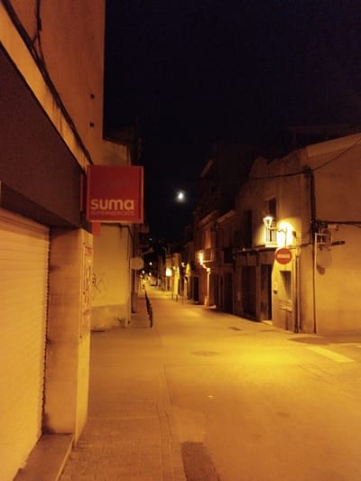 The image depicts a street scene at night in Rubí, Spain. The street is illuminated by yellow streetlights, casting long shadows. On the left is a building with a closed storefront displaying a red "Suma Supermercats" sign. The street extends into the distance, bordered by buildings with balconies and various architectural details. A "no entry" sign is visible on one of the buildings on the right. A faint moon is present in the dark sky above the street. The street surface is a mix of pavement and brickwork. The scene is deserted with no people visible.