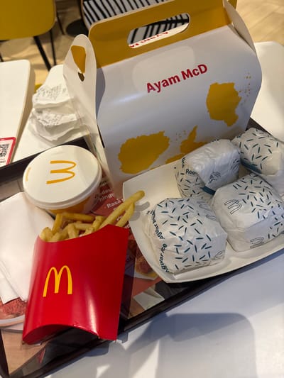 The image depicts a McDonald's meal on a tray, likely in a restaurant setting. The meal includes:

*   A box labeled "Ayam McD," which appears to be a chicken product.
*   Several McDonald's burgers wrapped in white paper with blue markings.
*   A red container of McDonald's French fries.
*   A white plastic cup, also with the McDonald's logo, likely containing a drink.

The food items are arranged in a somewhat haphazard manner on the tray. The tray itself appears to be black and is placed on a surface that might be a table. The setting is possibly a McDonald's restaurant in Bencongan Indah, Indonesia, given the "Ayam McD" product, a local menu item. Other details include a chair and some scattered paper waste.