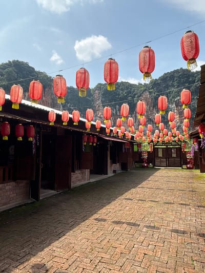 This image depicts the exterior of a building complex, possibly a tourist attraction, located in Ipoh, Malaysia, near a karst limestone hill. The scene is characterized by numerous red Chinese lanterns strung across wires, creating a festive atmosphere. The architecture features traditional wooden structures with brown facades and tiled roofs, complemented by brick walls at the base. A pathway made of reddish-brown brick pavers, laid in a herringbone pattern, leads through the courtyard, with some areas showing patches of moss. In the background, the imposing, rugged terrain of a limestone hill rises, covered with lush green vegetation. The sky is a clear blue with scattered white clouds. Several figures, possibly statues or decorative elements, are visible near an ornate wooden gate at the far end of the courtyard. The overall impression is that of a cultural heritage site or a recreational area designed to evoke traditional Asian aesthetics.