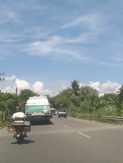 The image depicts a sunny day in Glee Gogo, Indonesia. A person is riding a motorcycle on a road, carrying various items on the back. In front of the motorcycle is a truck filled with white sacks of fertilizer. The truck has text on its side that reads "PUPUK NITREA". The road is asphalt and lined with yellow markings. The scene is surrounded by lush green trees and vegetation, with a bright blue sky dotted with fluffy white clouds. The time of day appears to be midday, given the bright sunlight and shadows. The overall impression is a typical day of transportation and commerce in a tropical region.