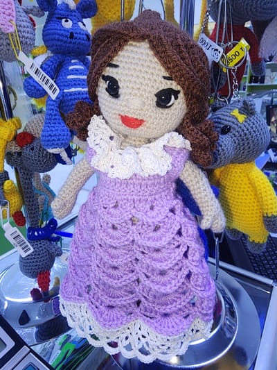 A crocheted doll with brown yarn hair and a light beige face is displayed in what appears to be a store or market stall. The doll is wearing a ruffled white collar and a lavender dress with intricate crochet patterns, trimmed at the hem with a cream-colored crocheted lace. The doll is positioned in the foreground, with other crocheted figures, including a blue dragon-like creature and a gray and yellow cat-like creature, visible behind it. Several price tags are attached to the items, with visible text including "0 L30", "000 T", and "1000". The background is blurred, suggesting a commercial setting with shelves and other merchandise. The overall scene depicts a display of handmade crocheted toys. The image was taken in Ushtobe, Kazakhstan.