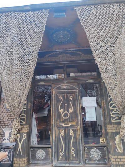 The image shows the entrance to a business in Alexandroupolis, Greece. The entrance is framed by two decorative nets and features a dark wooden door with ornate carvings. Above the door, there is a window with more netting visible behind it. A sign is affixed to the door that reads "ΔΕΥΤΕΡΑ 29 ΙΟΥΝΙΟΥ ΚΛΕΙΣΤΑ" which translates to "Monday, June 29th, Closed". Below this, there is another sign with a schedule of operating hours and a "TIMOKATALOGOS" which means "Price list". A "WARNING VIDEO SURVEILLANCE" sticker is present on the lower right of the door. The facade of the building is decorated with brickwork and painted designs, including images of scissors on the left and a comb on the right. The overall impression is of a business with a unique, perhaps rustic or artisanal, aesthetic.