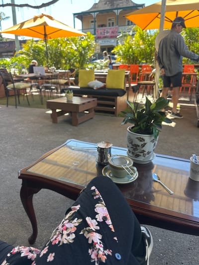 The image depicts an outdoor seating area at a cafe, likely in Bellingen, Australia, given the provided context. In the foreground, a person's legs are visible, wearing black pants with a floral pattern and sneakers. They are seated at a dark wood coffee table with a glass top. On the table, there is a teacup with a metal strainer, a small metal pitcher, a spoon, and a potted plant in a decorative white pot with brown floral designs. In the background, there are more tables and chairs, some with yellow cushions, under orange umbrellas. A building with a balcony and several windows is visible in the distance, along with lush green foliage. People are seen in the background, including a man in a gray shirt and shorts, and another person seated at a table. The overall atmosphere suggests a relaxed daytime setting at an outdoor cafe.