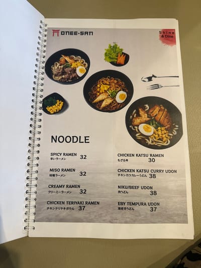 The image captures an open, spiral-bound menu from a restaurant named "ONEE-SAN" and "Shine & Dine," resting on a light brown, textured surface, likely a table. The menu page showcases a variety of noodle dishes under the heading "NOODLE," accompanied by appetizing photographs and prices.
The subjects are various food items, primarily noodle bowls such as ramen and udon. Objects include four distinct bowls of noodles with diverse toppings, a small side dish of greens and sliced carrots, and another small dish containing corn and spinach. A graphic of a spoon and fork is visible at the top of the page.
The activity depicted is the presentation of a food menu. The scene suggests a dining establishment in Kecamatan Cisaat, Indonesia.
Notable details include the clear, vibrant images of the dishes, showing ingredients like chicken katsu, sliced beef, half-boiled eggs, sweet corn, and various garnishes. The menu is bilingual, featuring both English and Japanese text for each item.
Visible text from the menu includes:
* Restaurant names: "ONEE-SAN," "Shine & Dine"
* Section title: "NOODLE"
* Menu items and prices:
* SPICY RAMEN (辛いラーメン) - 32
* MISO RAMEN (味噌ラーメン) - 32
* CREAMY RAMEN (クリーミーラーメン) - 32
* CHICKEN TERIYAKI RAMEN (チキンテリヤキボウル) - 37
* CHICKEN KATSU RAMEN (もざる丼) - 30
* CHICKEN KATSU CURRY UDON (チキンカツカレーうどん) - 38
* NIKU/BEEF UDON (肉うどん) - 38
* EBY TEMPURA UDON (海老天うどん) - 37