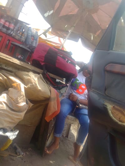 This image depicts an outdoor, informal commercial setting in the city of Mista Ali, Nigeria, likely a roadside stall under bright daylight. A female individual is seated on a light-colored plastic chair, positioned to the right of the frame. She wears a white t-shirt with a colorful graphic (partially obscured, appearing as "KISS B" with green, purple, pink, and red lettering), blue jeans rolled at the ankles, and is barefoot on one visible foot while the other is partially obscured near a sandal. She has a watch on her left wrist, rings on her fingers, and a black handbag with a gold chain strap on her lap. Her right arm is raised, with sunglasses possibly on her head or eyes.
To her left, a red and black duffel bag rests on a wooden surface. Adjacent to the bag are several beige tarps or sacks, with an orange and brown cloth draped over them. Behind this setup, various beverages are visible on a red shelf or rack, including Coca-Cola bottles, "TORA" branded bottles, and clear water bottles, alongside a black plastic basin. The area is shaded by two large market umbrellas, one reddish-brown/tan and another striped with yellow and red/orange. On the far right, the open front passenger door of a dark-colored vehicle is visible, revealing a worn interior panel. The ground appears to be dirt or dusty earth.