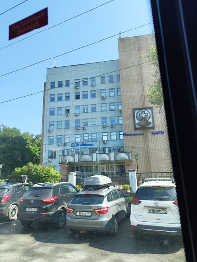 The image shows a view of the exterior of a medical facility, likely a clinic or hospital, in Zhukovsky, Russia. The building is a multi-story, light-blue concrete structure with numerous windows and visible air conditioning units. On the facade, there is a blue sign with Russian text, the larger part reading "АЛИМЕД" and a smaller line below it "МЕДИЦИНСКИЙ ЛЕЧЕБНО-ДИАГНОСТИЧЕСКИЙ ЦЕНТР 2 ЭТАЖ." To the right, on a brick-clad section of the building, a logo depicting a medical cross above a stylized bowl is visible, with the Russian words "Поликлиника ЦАГИ" below it. Several cars are parked in front of the building, including a black Hyundai Creta, a silver Lada Vesta, and a white Nissan X-Trail. A red sign with white text reading "ЗАПАСНЫЙ ВЫХОД" (Emergency Exit) is suspended in the sky, likely attached to overhead wires. The sky is clear and blue, and green trees are visible behind the building. The image is taken from inside a vehicle, suggested by the tinted window frame on the right side of the frame and the reflections on the glass.